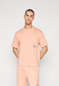Tricou cu mânecă scurtă, de culoare roz pal, realizat din bumbac moale, având o mică patch cu logo alb și negru pe partea stângă a pieptului.