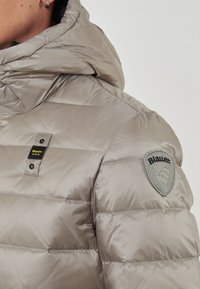 Giacca imbottita beige con colletto alto, etichetta di tessuto Blauer U.S.A. sul petto e logo ricamato Blauer sulla manica superiore.