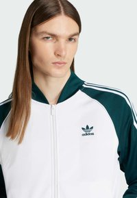 Bílá zipová sportovní bunda s zelenými akcenty na ramenou, se třemi bílými pruhy a malým zeleným logem Adidas na hrudi.