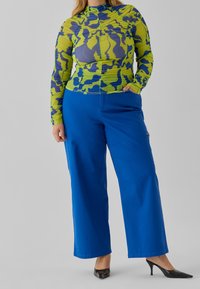 Maglietta a maniche lunghe in un vivace motivo camouflage verde e blu, abbinata a pantaloni larghi blu e scarpe con tacco alto nere.