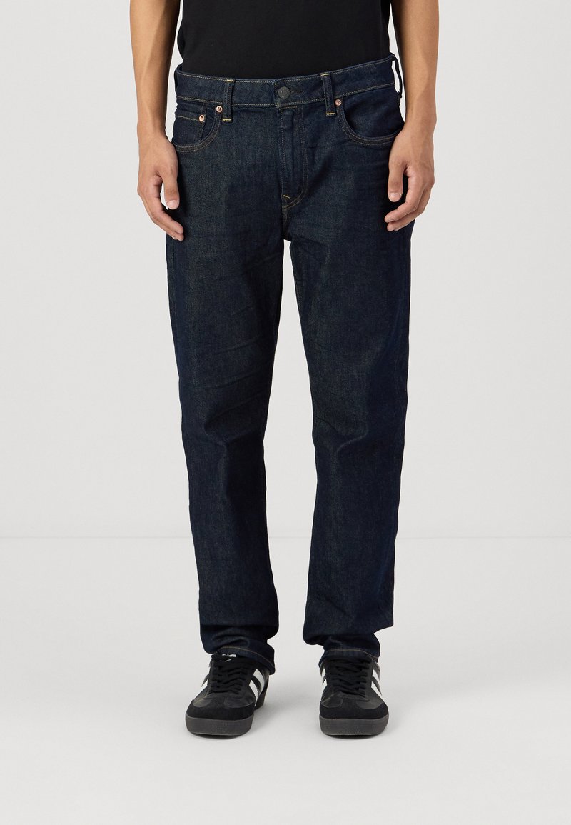 Tumši zili džinsi ar taisnu piegriezumu, kas satur piecas kabatas, varščaus un redzamām šuvēm. Valkāti ar melniem sporta apaviem.