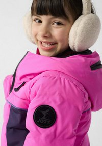 Chaqueta acolchada rosa con un panel azul marino, bolsillos con cremallera y un parche redondo con logo negro en la manga. La modelo lleva orejeras blancas suaves.