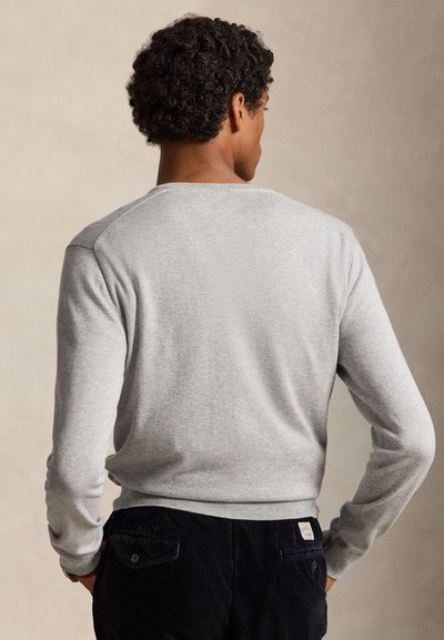 Man met krullend donker haar, gekleed in een lichtgrijze trui met lange mouwen en een zwarte broek, staand met handen in de achterzakken, met de rug naar de kijker toe.