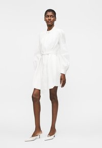 Femme portant une mini-robe blanche à manches longues boutonnée avec une ceinture à la taille et des talons blancs pointus, debout devant un fond uni.