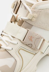 Beige högskor med texturerat material, justerbart spänne, elastiska skosnören och logotypdetalj. Har en kombination av släta och mesh-ytor.