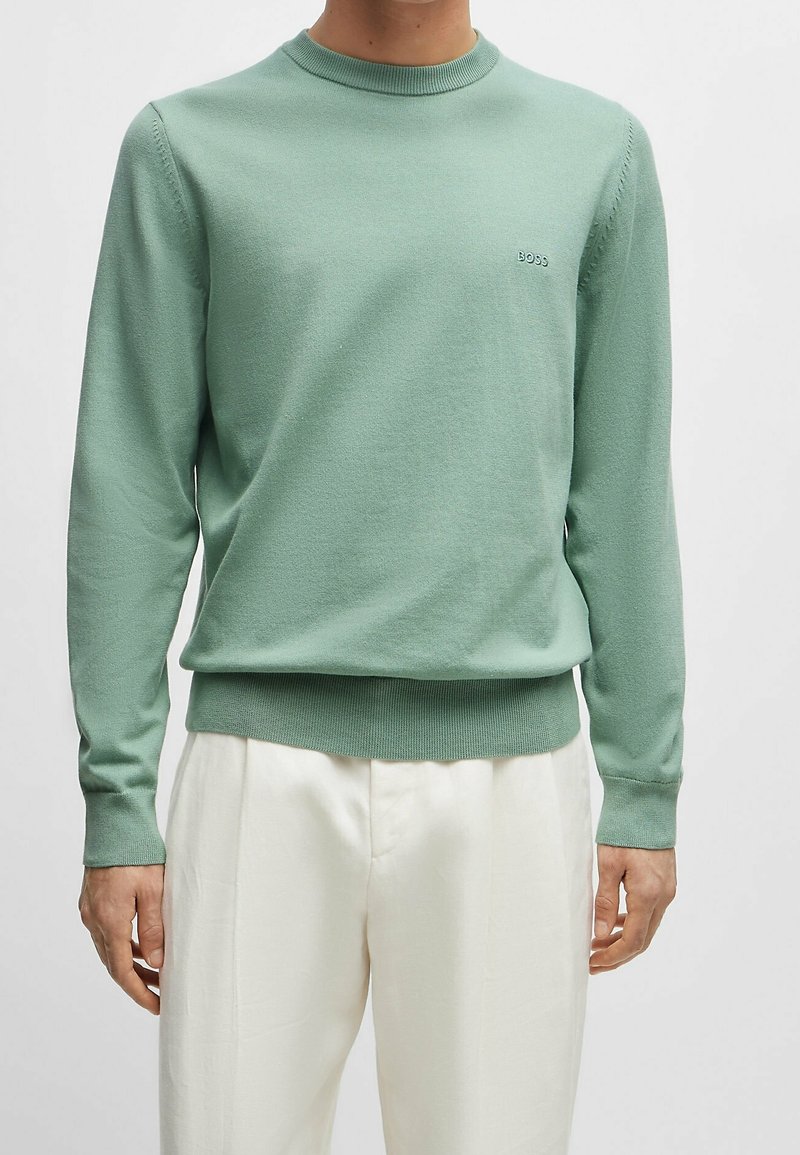 Homme portant un pull vert clair à manches longues avec le logo "BOSS" sur la poitrine et un pantalon blanc, debout devant un arrière-plan uni.