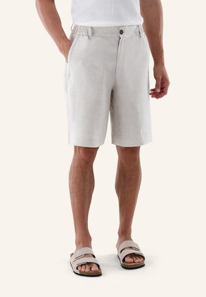 Mann trägt beige knielange Shorts mit elastischem Bund, weißes strukturiertes Hemd und beige Sandalen, steht vor weißem Hintergrund.