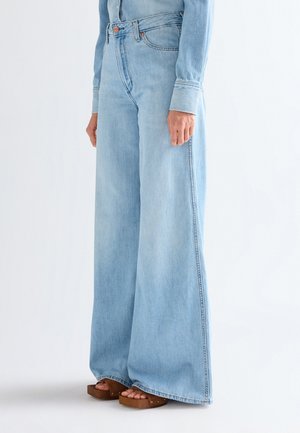 Osoba ubrana w jasnoniebieskie, szerokie dżinsy z wysokim stanem oraz masywne brązowe sandały na platformie, stoi z rękami swobodnie opuszczonymi wzdłuż ciała.