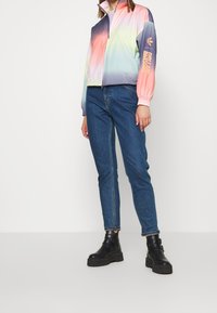 Färgglad vindjacka i pastellfärger av rosa, grön och lila med hög krage; kombinerad med blå denimjeans och svarta klumpiga stövlar.