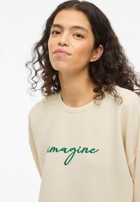 Beige oversized genser med korte ermer, med grønn brodert tekst "imagine" over brystet. Mykt bomullsmateriale, avslappet passform.