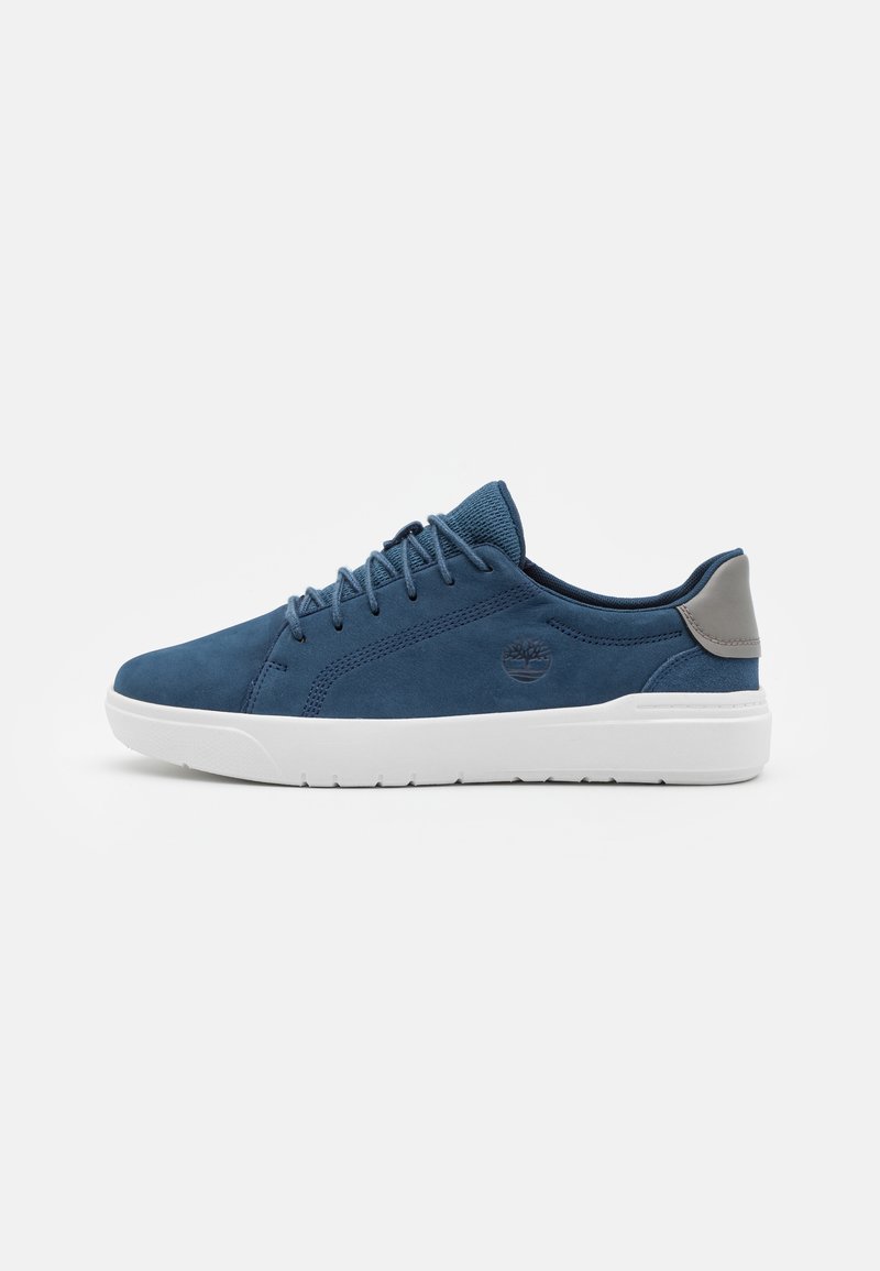 Zapatilla de ante azul con suela de goma blanca. Presenta un diseño con cordones, un collar acolchado y un logotipo sutil en el lateral. Material texturizado.