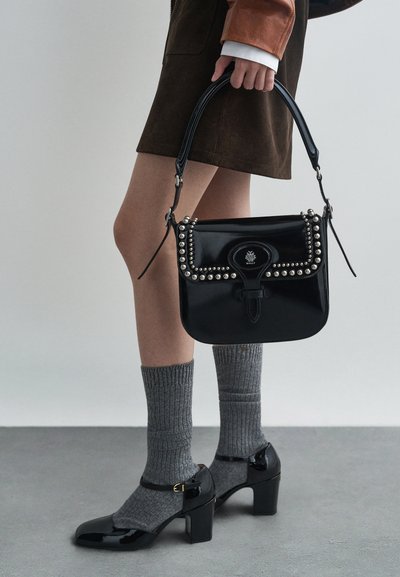 Borsa a mano in vernice nera con borchie metalliche e una patta decorativa. Abbinata a calze grigie a coste e scarpe con tacco a blocco in vernice nera.