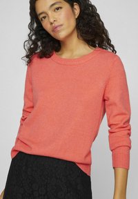 Korallfarget genser med rund hals og lange ermer, laget av mykt strikkemateriale; har ribbestrikkede mansjetter og en avslappet passform.