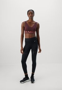 Athletische Frau in einem kastanienbraunen Sport-BH und schwarzen Nike Leggings, mit schwarzen Socken und schwarzen Turnschuhen, vor einem schlichten grauen Hintergrund.