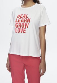 Camiseta de algodón blanca con texto rojo en negrita: "SANAR, APRENDER, CRECER, AMAR." Mangas cortas, cuello redondo y corte relajado. Combinada con pantalones rosas.