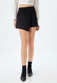 Schwarze Wickelhose aus glattem Stoff, mit seitlichem Knopfverschluss und zwei vorderen Taschen. Getragen mit schwarzen Socken und spitzen Absätzen.