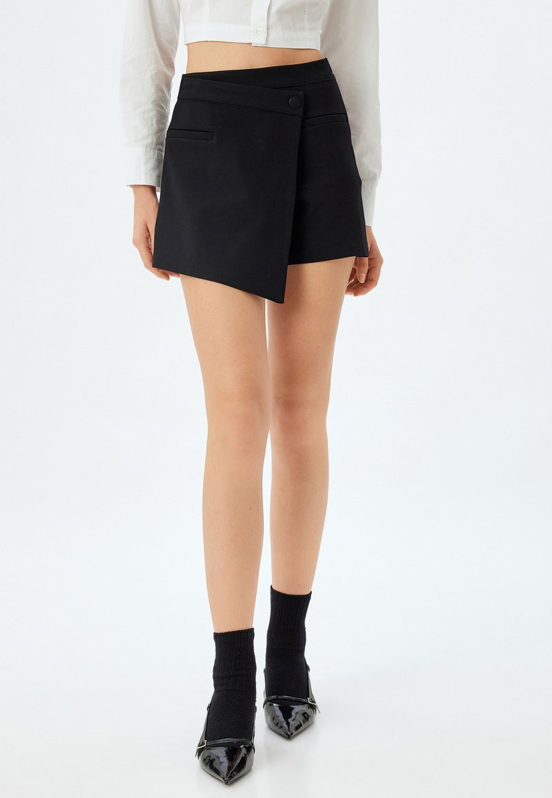Schwarze Wickelhose aus glattem Stoff, mit seitlichem Knopfverschluss und zwei vorderen Taschen. Getragen mit schwarzen Socken und spitzen Absätzen.