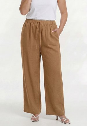 Kvinde iført løstsiddende brune bukser med høj talje og snøre samt hvide hælsandaler, hånd i lommen, stående på hvid baggrund.