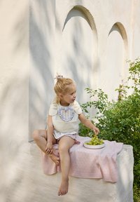 Barn i en cremet top med en lavendel volang, der sidder på et lys rosa tæppe; holder grønne druer på en hvid tallerken, med grønne planter i nærheden.