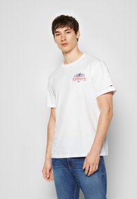 Hvid bomulds T-shirt med korte ærmer, med et farverigt "TOMMY'S" logo og et lille mærke på venstre ærme. Tætsiddende design.