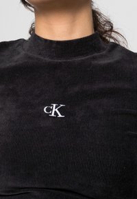 Frau trägt ein schwarzes, geripptes Oberteil mit hochgeschlossenem Kragen und einem weißen, gestickten "cK"-Logo auf der Brust.