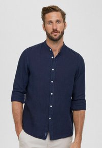 Camicia di lino blu navy con colletto alto, maniche arrotolate e bottoni bianchi, caratterizzata da una sottile trama e un'etichetta cucita sull'orlo.