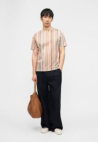 Νεαρός άνδρας που στέκεται με το ένα χέρι στην τσέπη, φοράει πουκάμισο με πορτοκαλί ρίγες και κοντά μανίκια, φαρδιά μαύρα παντελόνια, λευκά αθλητικά παπούτσια, κρατώντας μια καφέ δερμάτινη τσάντα.