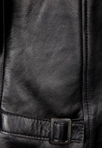 Gros plan sur du cuir noir texturé avec des coutures et une petite boucle métallique rectangulaire.