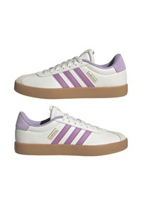 Zapatos de cuero blancos con tres rayas moradas, una suela de goma beige y acentos morados acolchados en el talón y la lengua. Detalle del logo en dorado.