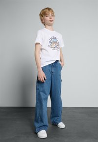 Bílý tričko s grafickým designem, kombinované s volnými modrými džíny. Model má na sobě bílé tenisky a stojí na šedém pozadí.