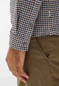 Camicia in flanella a quadri blu e marroni, con polsini a bottone. Abbinata a pantaloni marroni a trama, fissati con un bottone a pressione.