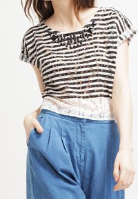 Stripet sort og hvid gennemsigtig bluse med blomstret blonde detaljer i kanten, med rund halsudskæring og dekorative sorte perle detaljer. Parret med blå plisserede bukser.