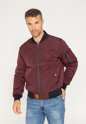Burgunderfarbene Bomberjacke mit Vorderreißverschluss, gerippten Bündchen und Saum. Verfügt über eine seitliche Reißverschlusstasche und ein oranges Etikett am Saum. Trägt man über einem grauen Shirt.