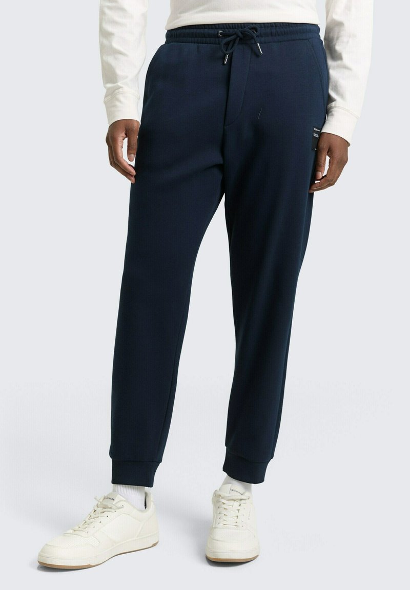 Mężczyzna noszący granatowe dresowe spodnie z elastycznymi mankietami, pas z sznurkiem, białe skarpetki i białe buty sportowe, stojący na jasnym tle.