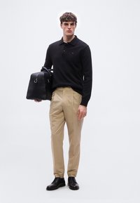 Άνδρας που φοράει μαύρο πόλο με μακριά μανίκια, μπεζ παντελόνι και μαύρα επίσημα παπούτσια, κρατώντας μαύρη δερμάτινη τσάντα duffel με φερμουάρ και λαβές.