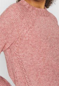 Pull en tricot rose avec encolure ronde, détail texturé sur le côté, poignets et ourlet côtelés, avec une apparence douce et duveteuse.