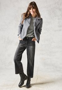 Veste en mélange de laine grise avec un col cranté, portée sur un pull gris texturé. Un pantalon large noir et des bottines noires complètent le look.