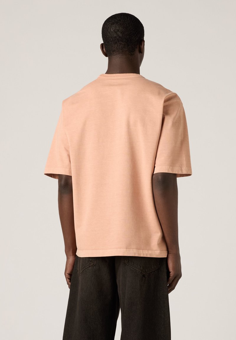 Mann trägt ein schlichtes pfirsichfarbenes Kurzarm-T-Shirt und lockere dunkle Hose, von hinten vor einem hellen neutralen Hintergrund gezeigt.