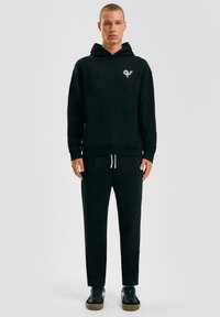 Zwarte hoodie met een voorzak en logo op de borst, gecombineerd met bijpassende zwarte trainingsbroek en zwarte sneakers met gumzolen.