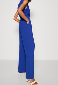 Blå ärmlös jumpsuit med avslappnad passform, sidofickor och vida ben. Tyget har en mjuk textur. Matching vit klacksandaler.