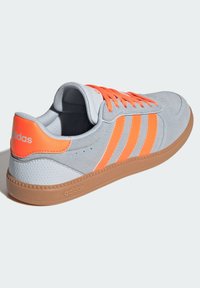 Šedá semišová teniska s oranžovými prvkami, tromi pruhmi a gumovou podrážkou. Má vypolstrovaný golier a kontrastné šnúrky.