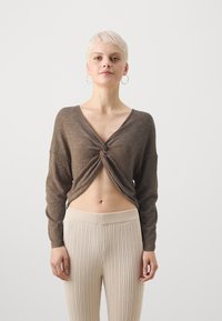 Top lungo a maniche lunghe marrone a costine con dettaglio annodato sul davanti, abbinato a pantaloni beige a costine a vita alta. Sfondo semplice.
