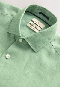 Chemise en lin verte avec col boutonné, tissu texturé et trois étiquettes indiquant la taille et les détails du matériau. Coupe régulière.