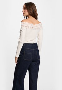 Cremfarbener schulterfreier Oberteil mit Spitzenbesatz, kombiniert mit hochtaillierten, dunkelblauen, weiten Jeans mit Gesäßtaschen und kontrastierenden Nähten.