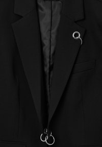 Il blazer nero presenta un design sartoriale con revers affilati, una fodera in satin e dettagli in metallo argentato, comprese le fedi alla chiusura.