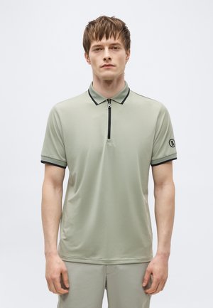 Bărbat tânăr purtând un tricou polo cu mâneci scurte de culoare verde deschis, cu margini negre, fermoar frontal și o insignă cu logo pe mâneca stângă.