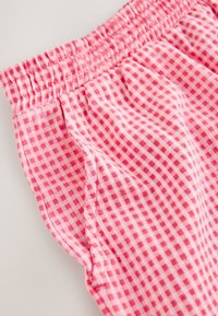 Tissu à carreaux roses et rouges avec taille élastique froncée et poche latérale visible sur un pantalon léger.