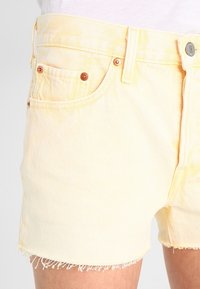 Pantalones cortos de mezclilla amarillo claro con dobladillo deshilachado, con bolsillos delanteros y detalles de botones de cobre. Diseño clásico de cinco bolsillos.