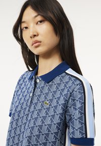Polo-paita sinisenä, jossa on geometrinen kuvio, tummansininen kaulus ja vaaleansiniset sivuraidat. Lacoste-logo rinnassa. Silkkinen pinta.
