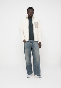 Fleece jacket in cream with a gray chest pocket, paired with a dark green T-shirt and light blue baggy jeans. Sneakers in white and blue.